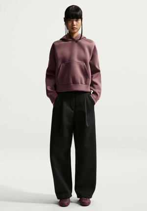 Ung kvinde stående iført en lilla hættetrøje, brede sorte bukser og matchende lilla sneakers med hænderne i lommerne mod en ensfarvet baggrund.