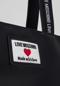 Schwarze Handtasche mit weißem Patch, der ein rotes Herz und den schwarzen Text „LOVE MOSCHINO Made with love“ zeigt, sowie einem markierten schwarzen Tragegurt.