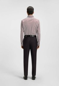 Homme de dos portant une chemise à rayures à manches longues, une ceinture noire, un pantalon foncé sur mesure et des chaussures de ville noires, devant un fond uni.