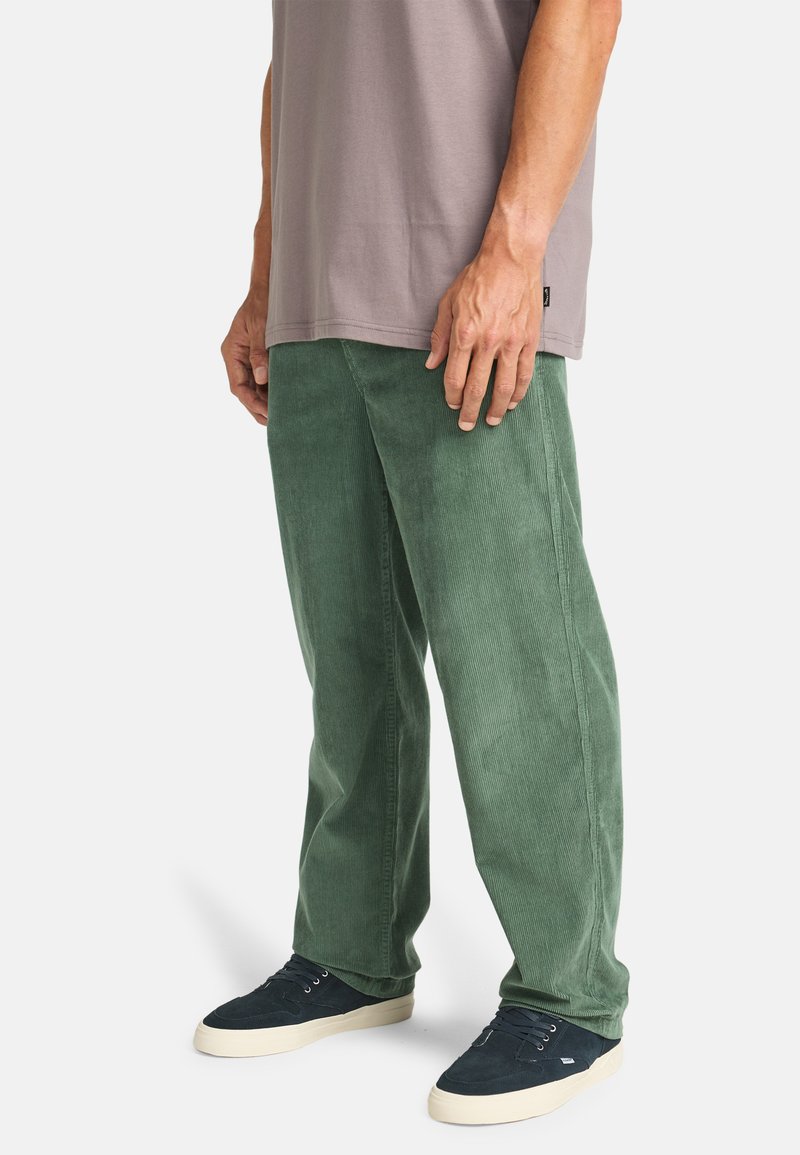 Green corduroy pants with a straight cut, featuring vertical ribbing and side pockets, paired with a gray t-shirt and dark shoes.