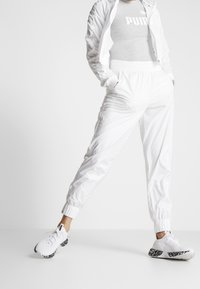 Bílá lehká sportovní bunda a bílé kalhoty s elastickými manžetami, doplněné bílými teniskami s texturovanou černou podrážkou.