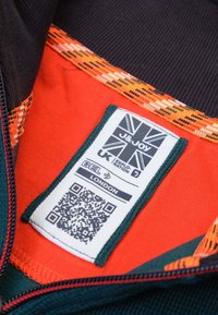 J&Joy-label op een zwarte stof met oranje binnenvoering en een patroon van oranje en rode afwerking. Bevat tekst, een QR-code en onderhoudsinstructies.