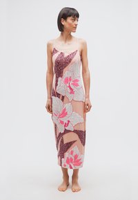 Γυναίκα με ροζ αμάνικο φόρεμα μέχρι τους αστραγάλους, διακοσμημένο με μεγάλα λουλουδένια παγέτες, στέκεται ξυπόλυτη μπροστά σε απλό λευκό φόντο.