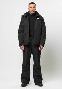 Schwarze wasserdichte Jacke mit Kapuze und Reißverschluss-Taschen, kombiniert mit passenden schwarzen Hosen. Glattes, strukturiertes Material und lockere Passform.