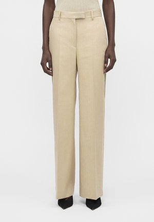 Μοντέλο φοράει μπεζ παντελόνες με φαρδιά πόδια, ζωνάκια και ψηλή μέση, σε συνδυασμό με μαύρες μυτερές γόβες.