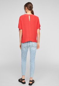 Blusa roja de manga corta, cierre en la parte de atrás con ojo de cerradura, combinada con jeans ajustados de color azul claro y sandalias negras de tiras. Textura de tela suave.