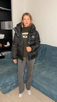 Giacca a vento nera con texture lucida, chiusura frontale con cerniera e collo ampio. Indossata sopra un maglione scuro e pantaloni grigi, con scarpe da ginnastica bianche.