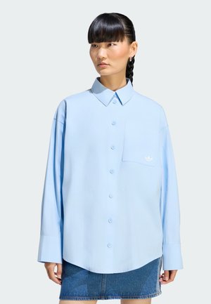Kvinde med flettet hår iført en lyseblå skjorte med knapper og denim nederdel, stående mod en neutral baggrund.