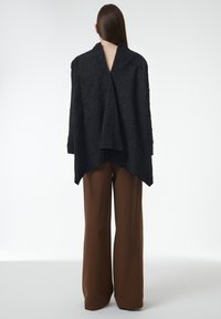 Prenda envuelta texturada en negro con escote en V, silueta fluida y dobladillo asimétrico, combinada con pantalones de pierna ancha marrones de talle alto.