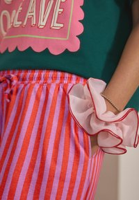 En person med grön skjorta med rosa text, röd och lila randiga byxor, och en genomskinlig rosa scrunchie på handleden med ett silverfärgat pärlhalsband.