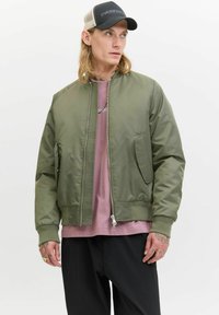 Maslinasto zeleni bomber jakna s glatkom, sjajnom teksturom, prednjim zipom i bočnim džepovima. Nosi se preko svijetlosmeđe majice.