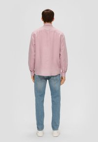 Rosa langärmliges Leinenhemd mit geknöpften Manschetten, getragen zu hellblauen Jeans und weißen Sneakers. Sichtbare Nähte und ein abgerundeter Saum.