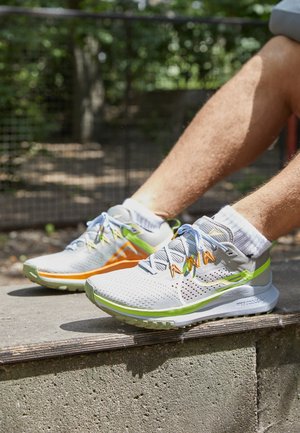 Lichte hardloopschoenen van grijze mesh met groene en oranje accenten. Ze hebben een dempende zool en reflecterende veters, ontworpen voor comfort.