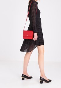 Sort kjole med polkaprikker og lange ærmer, parret med en rød crossbody taske. Sorte hæle med guldaccent. Gulvlængde, flydende stof.