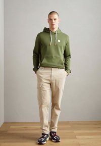 Oliven grønn hettegenser med snor, kengurulomme, kombinert med beige stripete cargo-bukser og fargerike joggesko. Bakgrunn av tregulv.