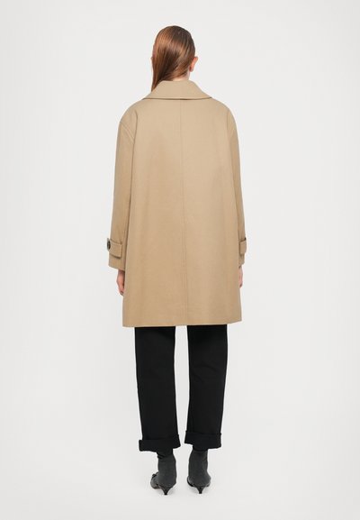 Cappotto beige con un ampio colletto, maniche lunghe e polsini abbottonati, abbinato a pantaloni neri. Il tessuto appare liscio e leggero.