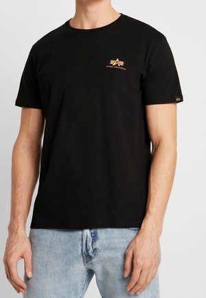 Hombre con camiseta negra de Alpha Industries con un pequeño logo naranja en el pecho y jeans azul claro frente a un fondo liso.
