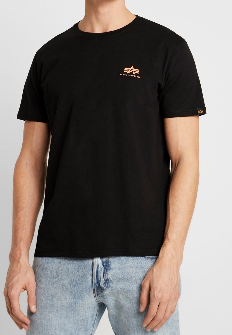 Muž oblečený v černém tričku Alpha Industries s malým oranžovým logem na prsou a světle modrých džínách na jednobarevném pozadí.