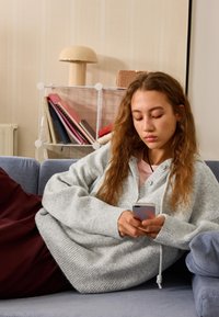Maglione grigio lavorato a maglia con vestibilità ampia e motivo testurizzato. Persona seduta su un divano blu, tiene in mano uno smartphone argento, circondata da libri.