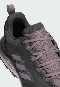 Zapato de senderismo gris y malva con una parte superior de malla, suela de goma y tres Rayas decorativas. Presenta una lengua texturizada y cordones duraderos.