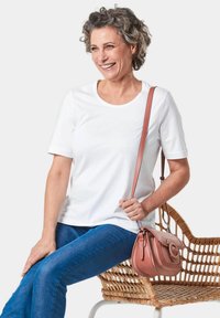 Witte korte mouwen t-shirt, blauwe jeans en een kleine roze crossbody tas. Vrouw zittend op een geweven stoel, lachend. Eenvoudig, casual ontwerp.