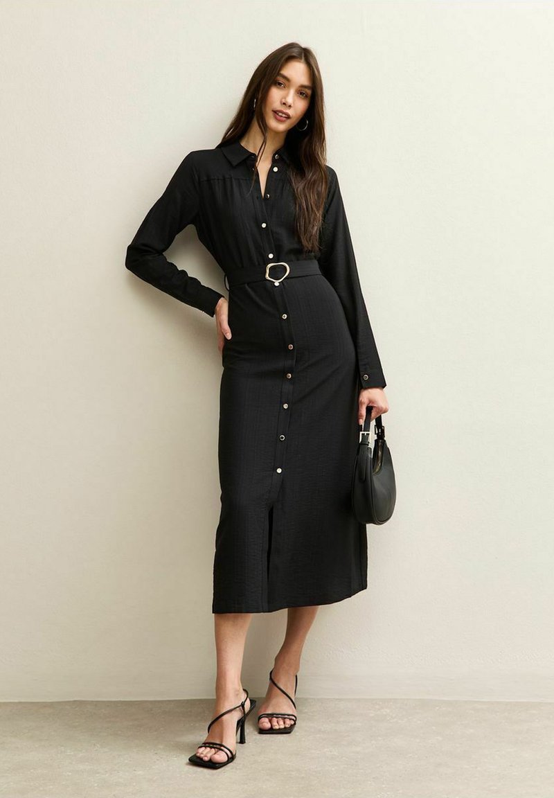 Abito camicia nero a maniche lunghe con bottoni, vita cintrata e spacco frontale. Accessoriato con una piccola borsa nera e sandali con lacci.