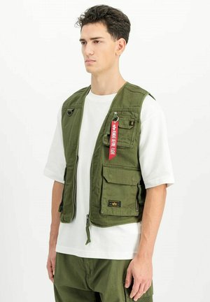 Giovane uomo che indossa un gilet utilitario verde oliva con multiple tasche e un'etichetta rossa "Remove Before Flight" sopra una maglietta bianca.