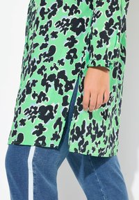 Tunique verde con motivo astratto nero, caratterizzata da spacco laterale e dettaglio con bottone sui polsini, abbinata a pantaloni di jeans blu con strisce chiare.