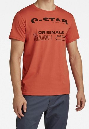 Mann trägt ein kurzärmeliges rostorangefarbenes T-Shirt mit schwarzem "G-STAR ORIGINALS RAW"-Schriftzug und graue Hose, steht mit entspannten Armen.