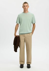 Pull vert menthe à manches courtes, pantalon beige et pull en maille marron foncé drapé sur le bras gauche. Le mannequin se tient devant un fond uni.