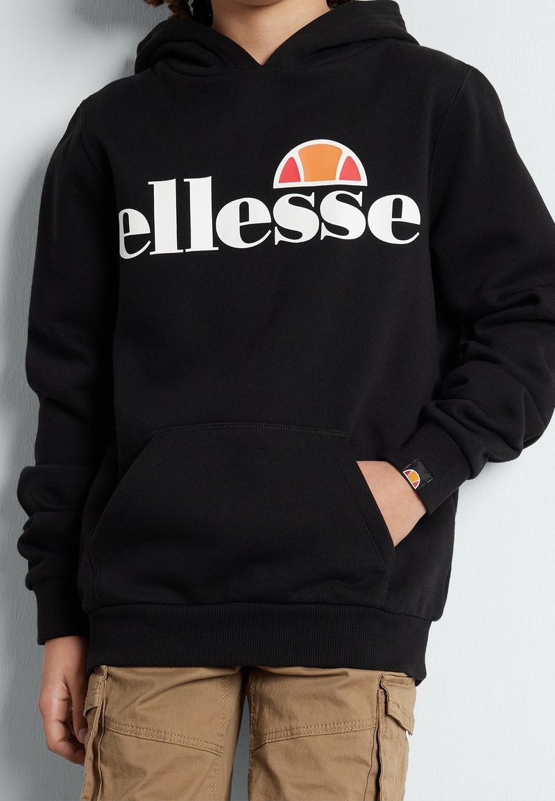 Enfant portant un sweat à capuche noir avec le logo "ellesse" blanc et orange, et un pantalon cargo beige, la main partiellement dans la poche du sweat.