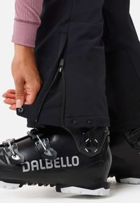 Zwarte skiboot met zilveren accenten en een textuur oppervlak, gecombineerd met zwarte buitenkleding met een rits en drukkers aan de zoom.