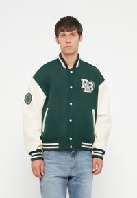 Grøn og creme-farvet varsityjakke med knaplukninger, ribstrikkede manchetter og et logo på brystet. Udstyret med sidelommer og stribede kanter.