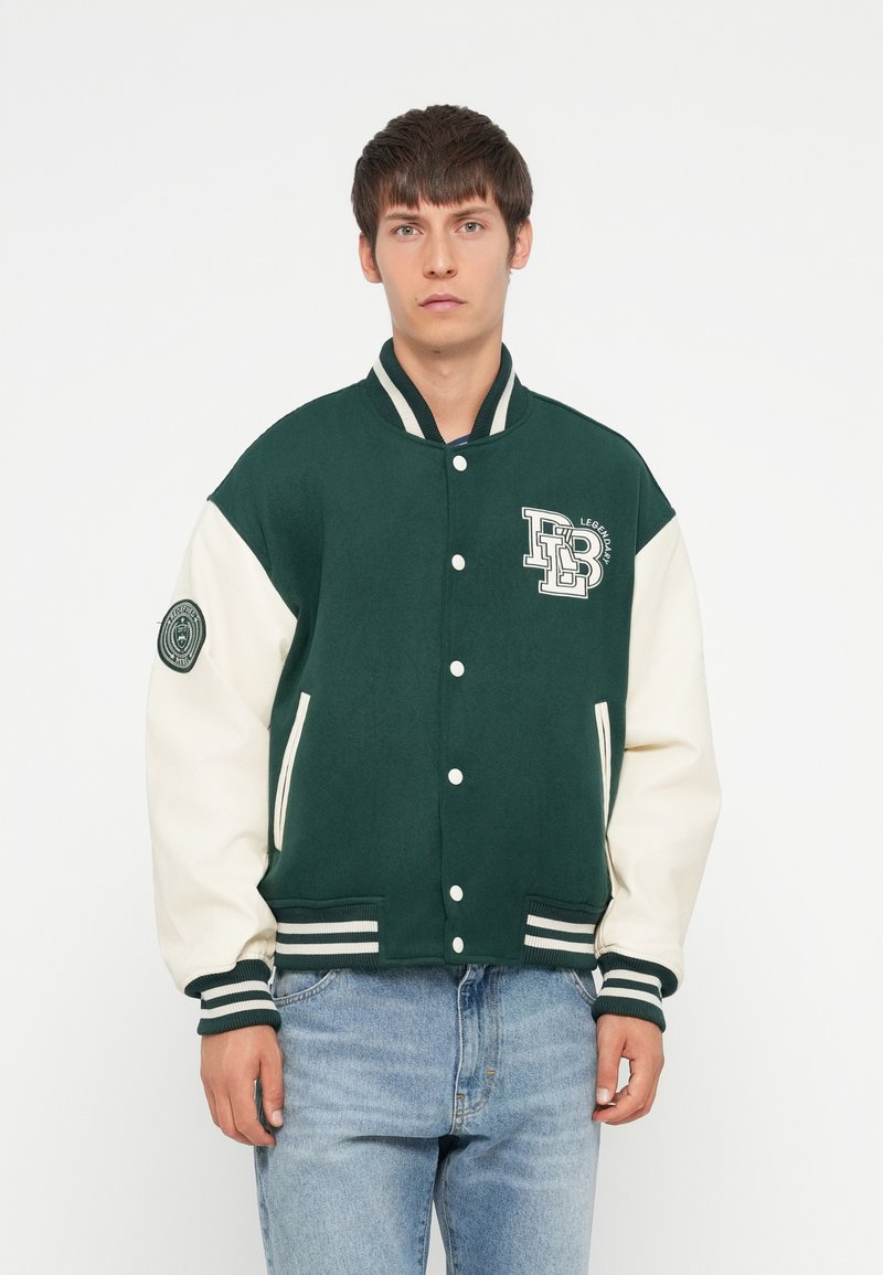 Grøn og creme-farvet varsityjakke med knaplukninger, ribstrikkede manchetter og et logo på brystet. Udstyret med sidelommer og stribede kanter.