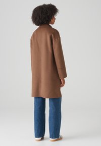 Brown wool coat with a relaxed fit, long sleeves, and minimal collar, paired with blue wide-leg jeans and white sneakers.
