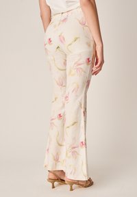 Frau trägt hoch taillierte, weit geschnittene cremefarbene Hosen mit rosa Blumenmuster und beige Sandalen mit Absatz, steht vor einem schlichten Hintergrund.