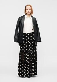 Γυναίκα που φοράει λευκό μπλουζάκι, μαύρο δερμάτινο μπουφάν περασμένο στους ώμους, μακριά μαύρη φούστα με λευκές πουά κουκίδες και μαύρα γάντια.