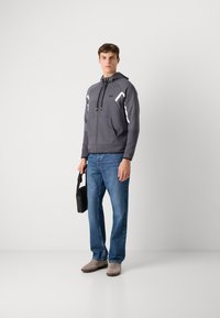 Pilkas zip-up džemperis su baltais tinkliniais akcentais, derinamas su mėlynomis tiesiomis kelnėmis ir šviesiai pilkais sportbačiais, laikantis juodą petinę rankinę.