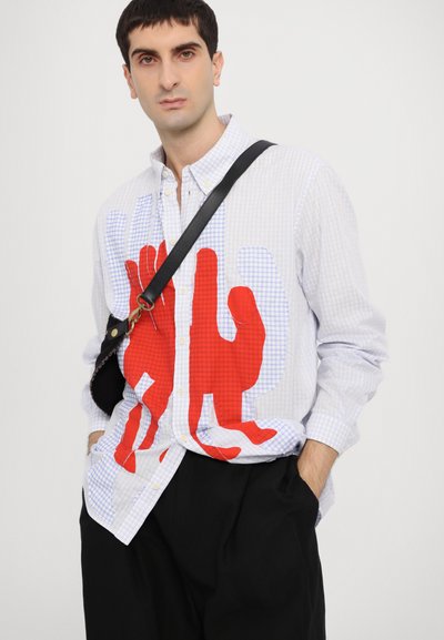 Camisa de botones con un patrón de cuadros blanco y azul, que presenta un gran gráfico de una mano roja. Combinada con pantalones negros y un bolso cruzado.