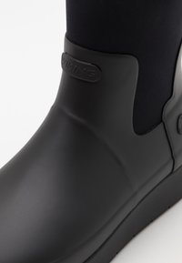 Melnas gumijas zābaki ar gludu virsmu, kas aprīkoti ar elastīgu augšdaļu, noapaļotu purngalu un ar zīmola siksniņu tuvu potītei, lai nodrošinātu drošu piegulšanu.