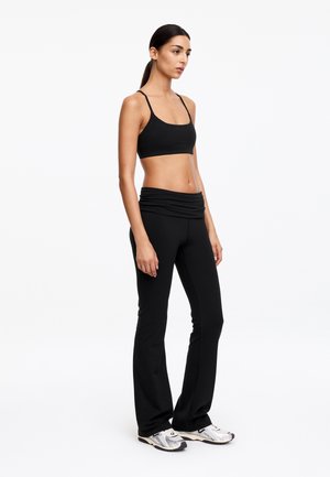 Femme aux cheveux foncés attachés, portant un soutien-gorge de sport noir, un pantalon de yoga noir et des chaussures de sport blanches, debout sur un fond blanc.