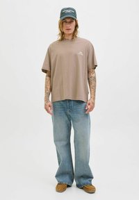 Lysebrun bomulds T-shirt med korte ærmer og et logo; parret med lyseblå denimjeans og brune slip-on sko. Mørkegrøn baseball cap.