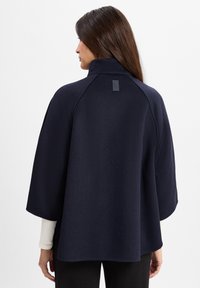 Granatowy wełniany poncz z wysokim kołnierzem, szerokimi rękawami i gładką teksturą. Posiada małą metkę z logo z tyłu przy szyi.