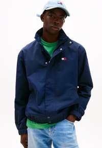 Marineblaue Windjacke mit Druckknöpfen, elastischen Bündchen und Logo-Aufnäher. Über einem grünen Hemd getragen, kombiniert mit hellblauen Jeans.