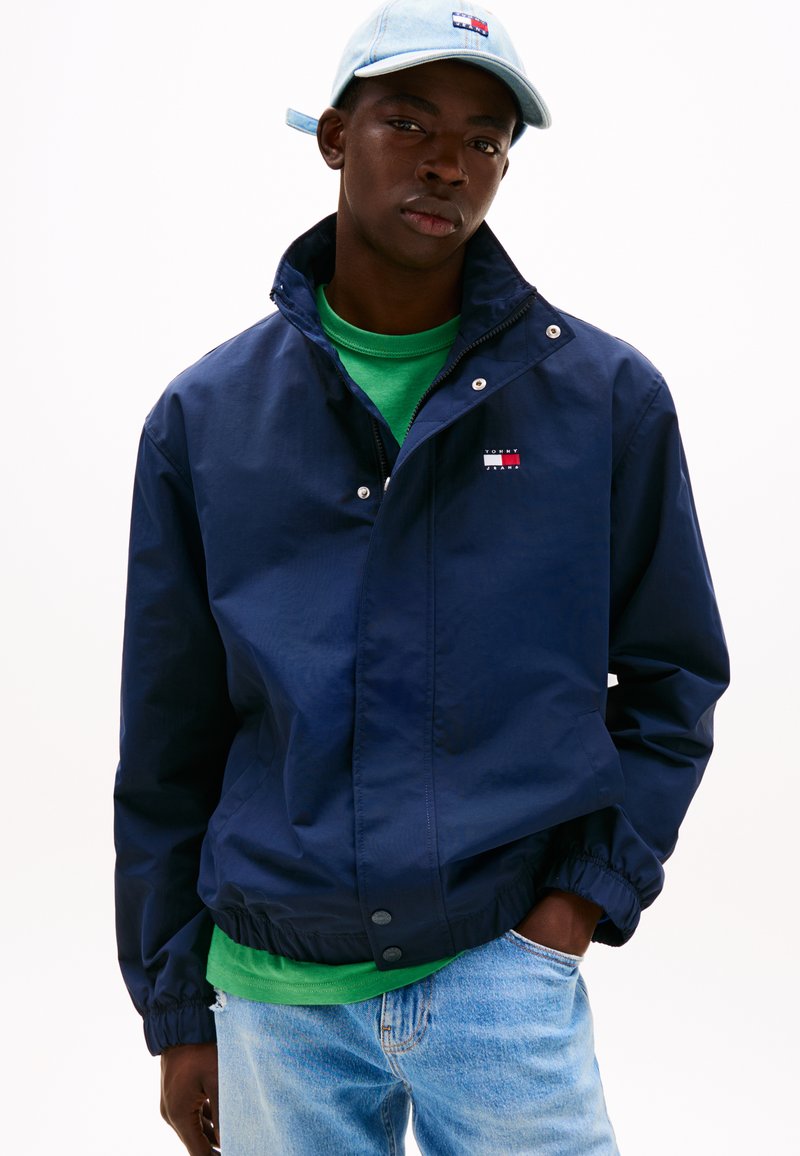 Marineblaue Windjacke mit Druckknöpfen, elastischen Bündchen und Logo-Aufnäher. Über einem grünen Hemd getragen, kombiniert mit hellblauen Jeans.