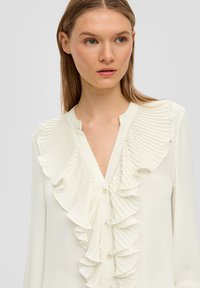 Weiße Bluse mit einem gerafften, plissierten Design an der Vorderseite, ausgestattet mit einer Knopfschließe und einer sanften, glatten Textur.