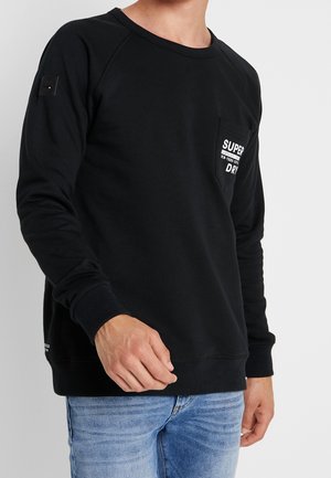 Mężczyzna w czarnej długiej bluzie z kieszenią z przodu, na której widnieje napis "Super Dry", w połączeniu z jasnoniebieskimi dżinsami na jednolitym tle.