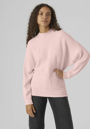 Suéter rosa acanalado con cuello alto, mangas largas y corte relajado, combinado con jeans oscuros. Textura suave y material blando.