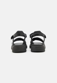 Sandales en cuir noir avec des sangles réglables, un semelle intérieure texturée et une semelle en caoutchouc épaisse, affichant un design minimaliste mais fonctionnel.