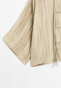 Vêtement beige léger en tissu texturé, à manches larges et longues lanières sur le devant pour la fermeture.